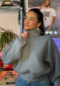 Jeune femme portant un pull gris zippé et des boucles d'oreilles en créole, debout en tenant le col, avec des plantes et une affiche en arrière-plan.