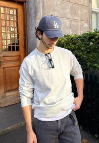 Sweatshirt cinza com mangas raglan, mostrando uma textura suave. Combinado com jeans escuros. Um boné azul-marinho com a letra 'LA' está usado. Óculos pendem do pescoço.