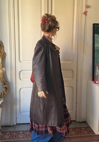 Καφέ δερμάτινο παλτό με εφαρμοστή γραμμή και με αναγκαία καλαίσθητη λαιμόκοψη, φορεμένο πάνω από μια καρό φούστα, συνδυασμένο με μπλε τζιν και μια κόκκινη τσάντα.