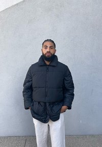 Μαύρο μπουφάν puffer με ψηλό γιακά, που διαθέτει φερμουάρ και κάθετες ραφές. Συνδυάζεται με ανοιχτόχρωμο παντελόνι.