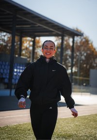 Casaco preto com fecho, capuz, punhos elásticos e logótipo da Nike; combinado com leggings pretas com logótipo da Adidas. Ambiente ao ar livre com céu limpo.