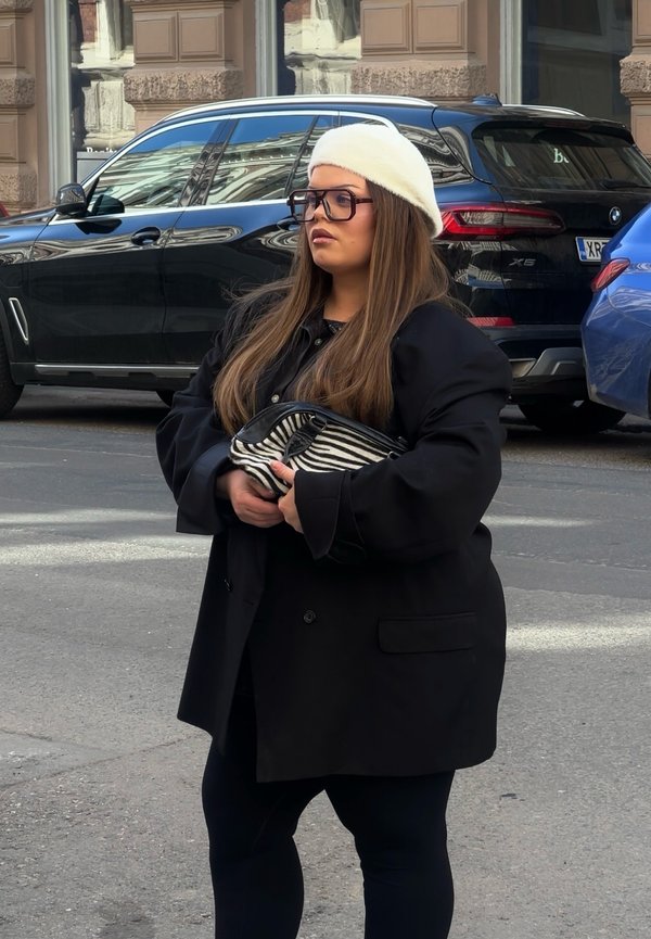 Kvinde iført en hvid baret, store briller og sort frakke holder en clutch med zebra-tryk og står på en byvej med parkerede biler bag sig.