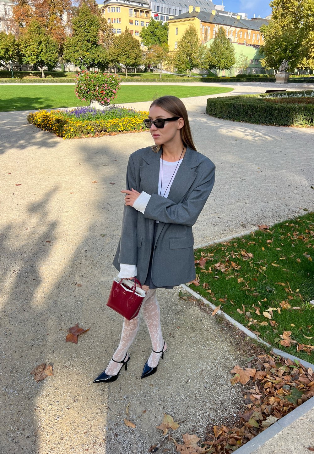 Grå oversized blazer med hvide manchetter, lagdelt over en hvid bluse. Sorte lakerede hæle, mønstrede strømper og en lille rød håndtaske.