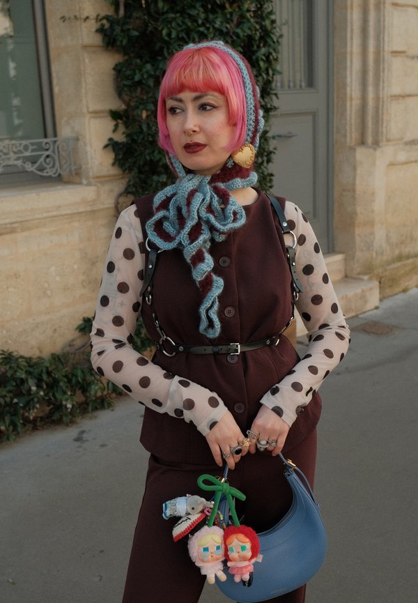 Personne aux cheveux roses et piercings au nez porte un haut à manches longues à pois, un gilet sombre, une écharpe en laine et tient un sac à main bleu contenant des peluches.