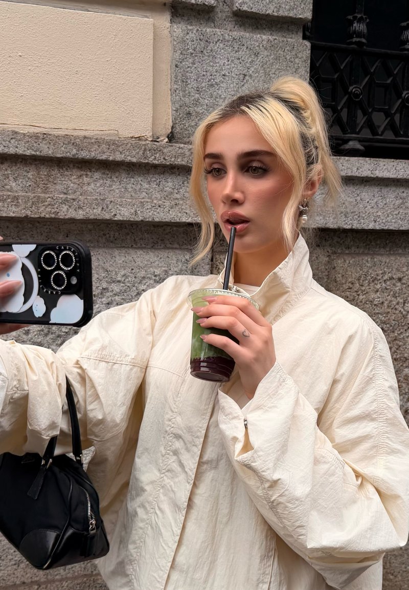 Young woman with blonde hair tied up, wearing a light jacket, drinking a green smoothie through a straw while taking a selfie outdoors.