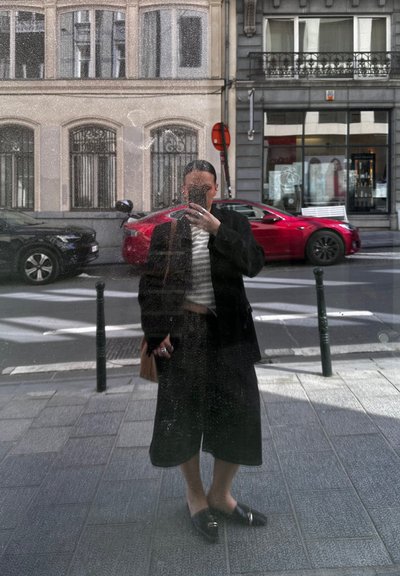 Abrigo negro de gran tamaño sobre una camiseta blanca a rayas, combinado con culottes negros y mocasines negros. Superficie de ventana reflectante, fondo urbano.