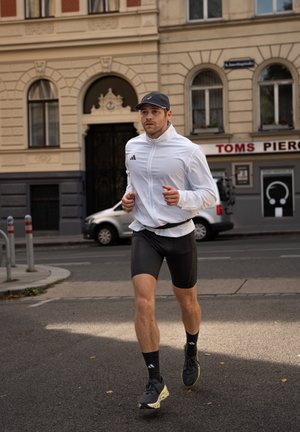 Runner in giacca bianca con zip e pantaloni corti neri, indossando un cappello nero e sneakers scure con suola chiara, su una strada cittadina.