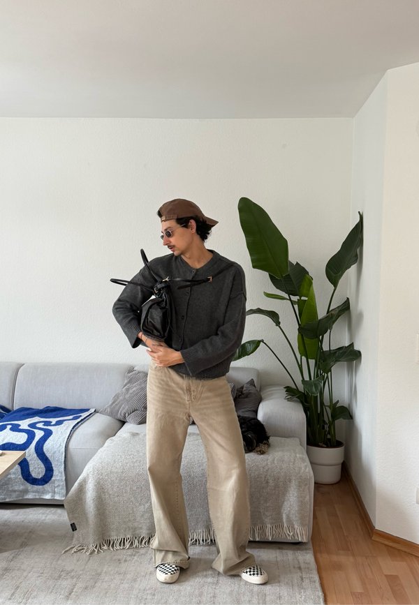 Junge Person mit Sonnenbrille und Kappe, die eine schwarze Tasche hält, in einem Wohnzimmer mit einer großen Pflanze und einem Sofa im Hintergrund.