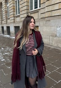Jeune femme aux longs cheveux blonds portant un manteau gris, une écharpe bordeaux, une chemise à rayures et une jupe plissée, debout sur un trottoir en ville.