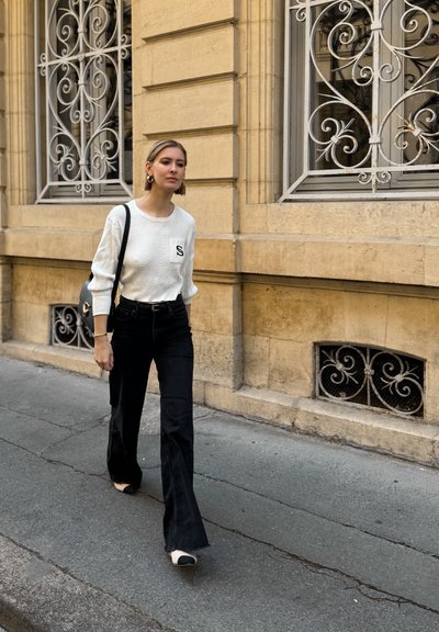 Suéter blanco de punto con un bolsillo y jeans negros acampanados. Bolsa negra con herrajes dorados, zapatos de color claro. Muro de piedra y forja en el fondo.