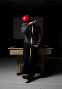 Un homme portant une casquette rouge s'appuie sur une queue de billard devant une table de billard dans une pièce faiblement éclairée.