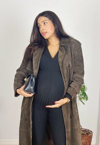 Femme enceinte en tenue noire et manteau marron tenant une pochette noire et caressant doucement son ventre, debout à l'intérieur près d'une plante.