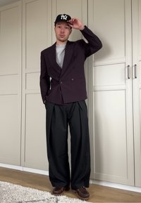 Doppelreihiger dunkelvioletter Blazer über einem grauen Hemd, kombiniert mit locker sitzenden schwarzen Hosen und braunen Schuhen, ergänzt durch eine schwarze Baseballkappe.