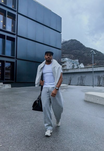 Hombre con pantalones deportivos grises, camiseta blanca, chaqueta gris sobre los hombros, gorra negra y zapatillas caminando al aire libre cerca de un edificio moderno con un fondo de montaña.