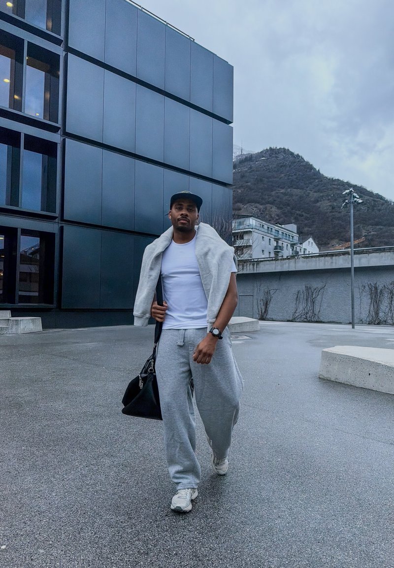 Homme en pantalon de survêtement gris, t-shirt blanc, veste grise portée sur les épaules, casquette noire et baskets, marchant à l'extérieur près d'un bâtiment moderne avec en arrière-plan des montagnes.