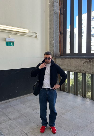 Chaqueta de punto negra, camiseta blanca, jeans azules y zapatillas rojas. Sostiene un bolso negro de hombro. Fondo de pared de concreto y una gran ventana.
