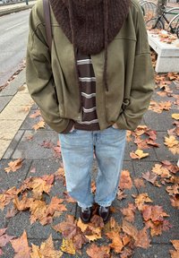 Veste oversize vert olive, écharpe tricotée marron, chemise rayée, jeans bleu clair et chaussures foncées sur un fond de feuilles tombées.