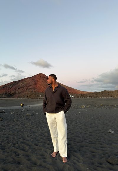 Hombre de pie descalzo en una playa de arena negra con colinas rocosas y cielo nublado de fondo durante el atardecer.
