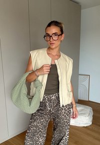 Μπεζ αμάνικο γιλέκο, ελαιόχρωμη μπλούζα, παντελόνια φαρδιά με λεοπαρδάλη στάμπα, πράσινη πλεγμένη τσάντα, μεγάλα γυαλιά και απλά κοσμήματα σε ουδέτερο φόντο.