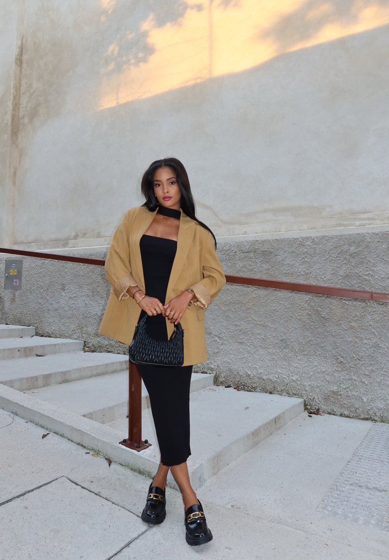 Μπεζ υπερμεγέθη σακάκι, μαύρο εφαρμοστό φόρεμα, μαύρη υφή τσάντα και μαύρα παπούτσια με χρυσές λεπτομέρειες; απαλές επιφάνειες, αντίθετα χρώματα.