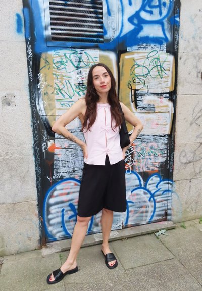 Top de color rosa claro, sin mangas, con lazo en el frente, pantalones cortos amplios negros y sandalias negras. El fondo presenta grafitis en una pared de concreto.