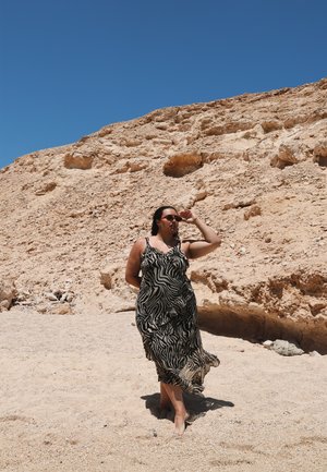 Vestido maxi de patrón de cebra en blanco y negro, con tirantes finos, tela fluida y un corte relajado; llevado con gafas de sol en una playa de arena.