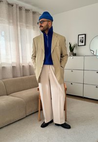 Blazer beige sur un col roulé bleu, pantalons larges blancs, chaussures noires et bonnet bleu. Intérieur avec des meubles de couleur claire.