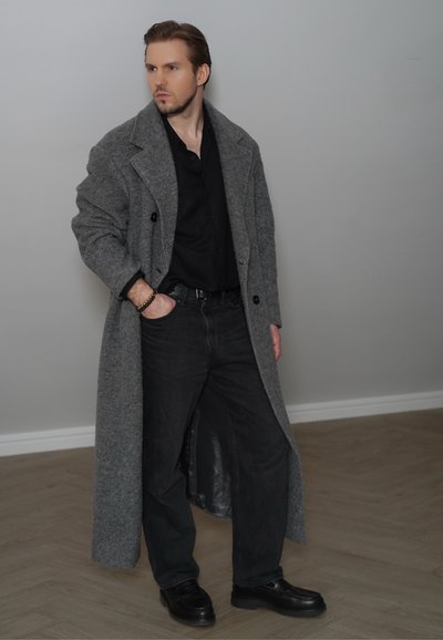 Abrigo de lana gris sobre una camisa negra, combinado con pantalones oscuros y mocasines negros; presenta un diseño de doble botonadura y tejido texturizado.