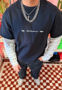 Personne portant des chemises à manches longues superposées noires et grises avec le texte "Quiksilver", des chaînes argentées avec une croix, un jean foncé et des baskets blanches sur un tapis à carreaux rouges.