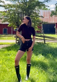 Schwarze Gummiregenstiefel, dunkler Oberteil und Jeansshorts, stehend im Gras mit Bäumen und einer roten Scheune im Hintergrund unter strahlendem Sonnenlicht.
