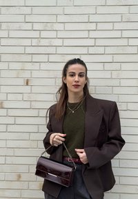 Femme aux longs cheveux foncés portant un blazer brun foncé, un haut vert olive, un pantalon gris, et tenant un sac à main brun foncé avec une bandoulière en chaîne.