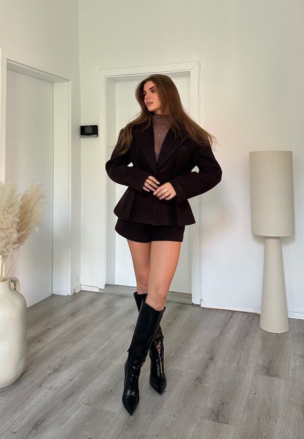 Femme debout à l'intérieur portant un blazer noir, un short assorti et des bottes noires hautes jusqu'aux genoux sur un parquet en bois clair.