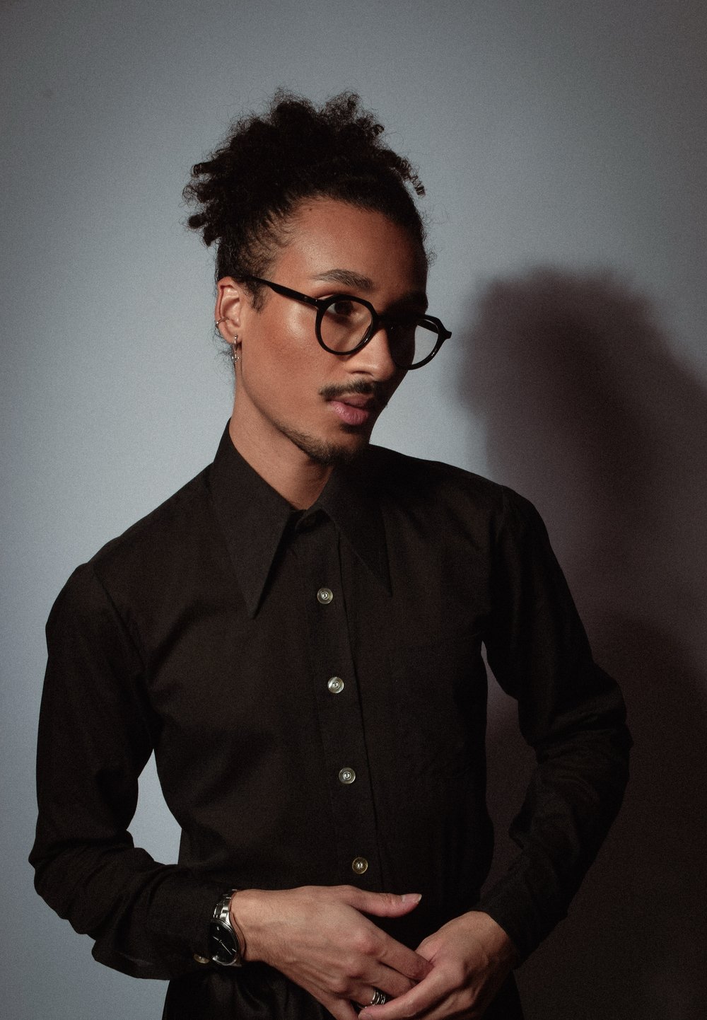 Chemise noire à boutons avec col pointu, grandes lunettes rondes et coiffure texturée, sur un fond neutre.