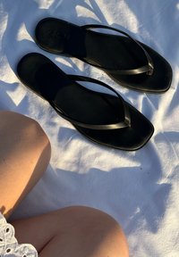 Chinelos pretos com tiras finas e sola plana, colocados sobre uma superfície branca. O calçado apresenta uma textura suave e um design minimalista.