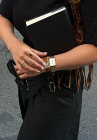 Svart notatbok holdt mot en arm med flere gullringer, et gullur på det ene håndleddet, og solbriller hvilende i hånden. Mørkt antrekk og en frynset veske synlig.