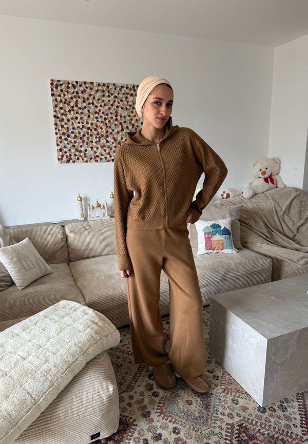 Mujer con conjunto de punto marrón y pañuelo beige en la cabeza, de pie en la sala de estar con sofá beige, alfombra con estampado, mesa de piedra gris y un oso de peluche en el sofá.