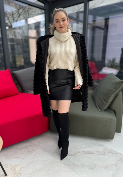 Suéter de tortuga crema, falda mini de cuero negra y botas negras hasta la rodilla. Abrigo de piel sintética negra. El fondo incluye asientos coloridos.
