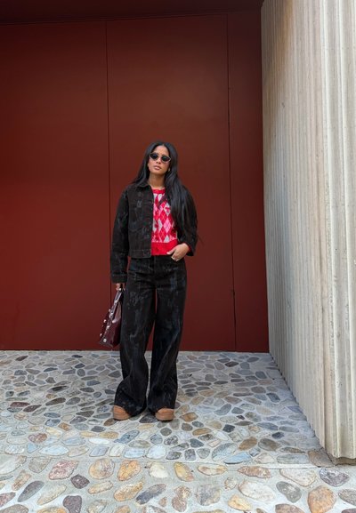 Mujer con cabello largo que lleva gafas de sol, suéter rojo con estampado debajo de chaqueta y pantalones negros, sosteniendo un bolso, de pie sobre un pavimento de piedra.