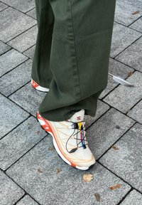 Beige sneaker with orange accents, reflective details, and black laces. The sole is chunky, featuring a textured pattern and logo.