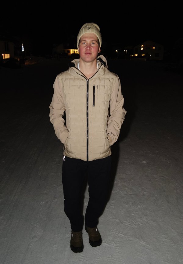 Mann trägt eine beige Winterjacke, beige Mütze, schwarze Hose und braune Stiefel, steht nachts im Schnee vor beleuchteten Häusern.