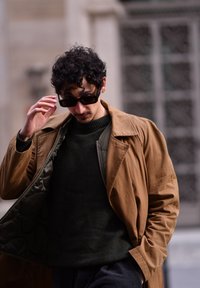 Hnedý trenčkot cez tmavo zelený sveter, doplnený tmavými slnečnými okuliarmi. Kabát má zips a prešívanú podšívku. Viditeľné kučeravé vlasy.