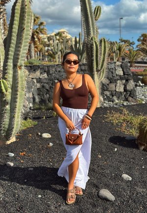 Mujer con gafas de sol, camiseta marrón, falda blanca y sandalias, de pie entre altos cactus y rocas volcánicas bajo un cielo parcialmente nublado.