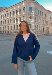 Sötétkék kardigán gombos elülső résszel, két zsebbel és széles ujjal, fehér inggel és világoskék farmerral párosítva. Városi háttér.