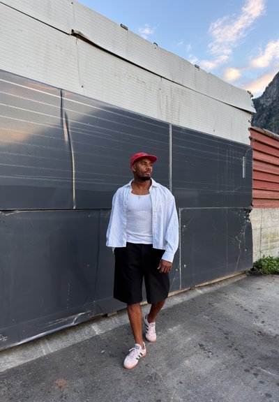 Hombre con gorra roja, camiseta de tirantes blanca, camisa a rayas y pantalones cortos negros caminando por la acera junto a una pared de metal ondulado.