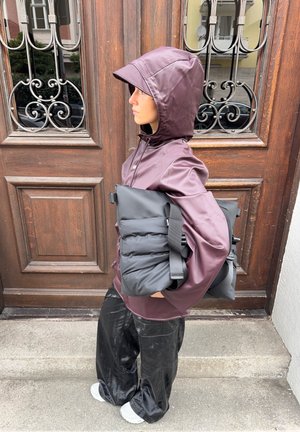 Chaqueta impermeable burdeos con capucha, bolsa acolchada negra con correas y pantalones negros brillantes. De pie contra una puerta de madera.