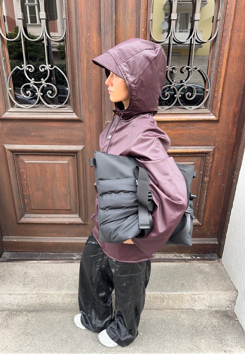 Giacca impermeabile color mogano con cappuccio, borsa imbottita nera con cinturini e pantaloni neri lucidi. In piedi contro una porta di legno.