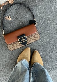 Bolsa transversal em couro castanho com estampa de logotipo e fecho preto, realçada por detalhes em dourado. Sapatos biqueira fina em camurça bege e jeans de ganga abaixo.
