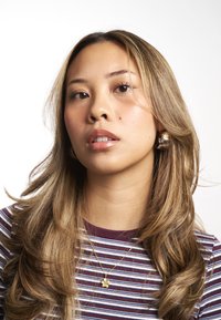 Portrait d'une personne aux cheveux longs, raides et ondulés de couleur brune, portant une chemise rayée maroon et blanche, un collier en fleurs dorées et des boucles d'oreilles en perles.
