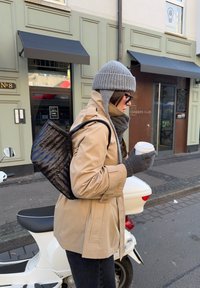 Czarny plecak z tkaniny, beżowy płaszcz trencz, szary dzianinowy kapelusz, szary szalik, szare rękawiczki, trzymający biały kubek z kawą, stojący obok skutera na ulicy.