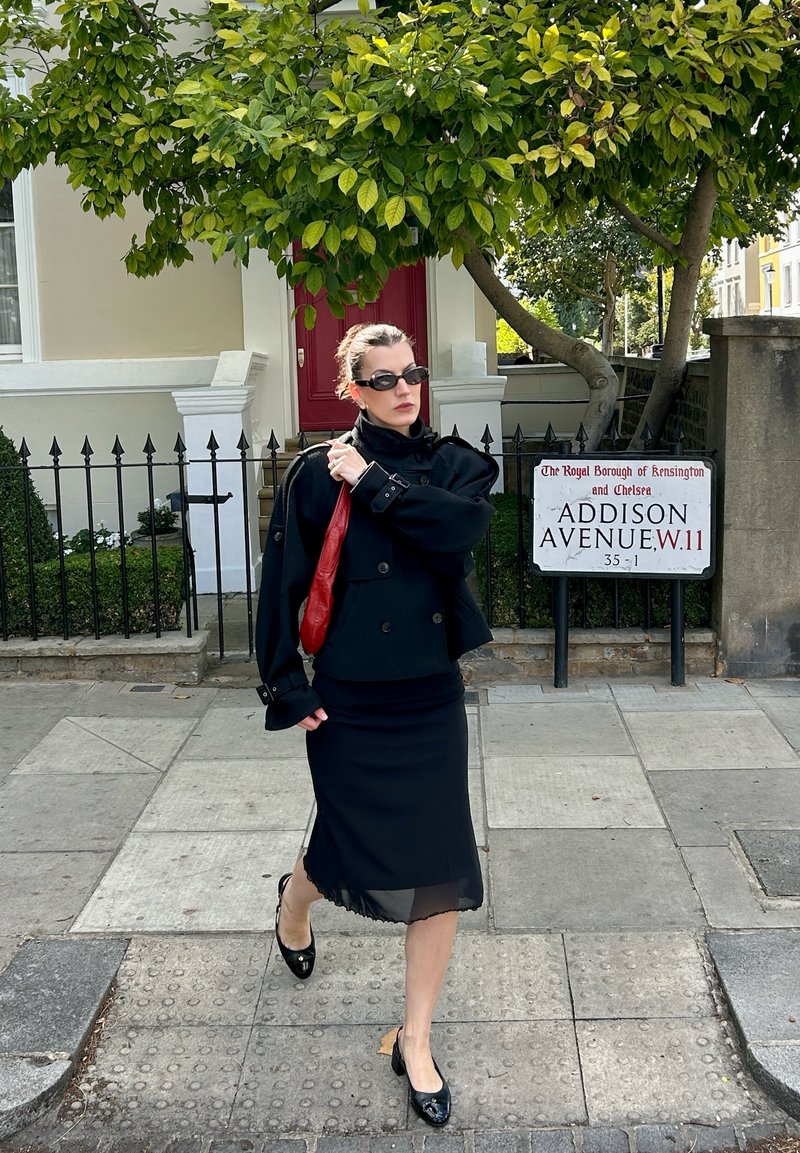 Woman in black coat and skirt walking on sidewalk near Addison Avenue street sign in Kensington, London, holding a red bag over her shoulder.